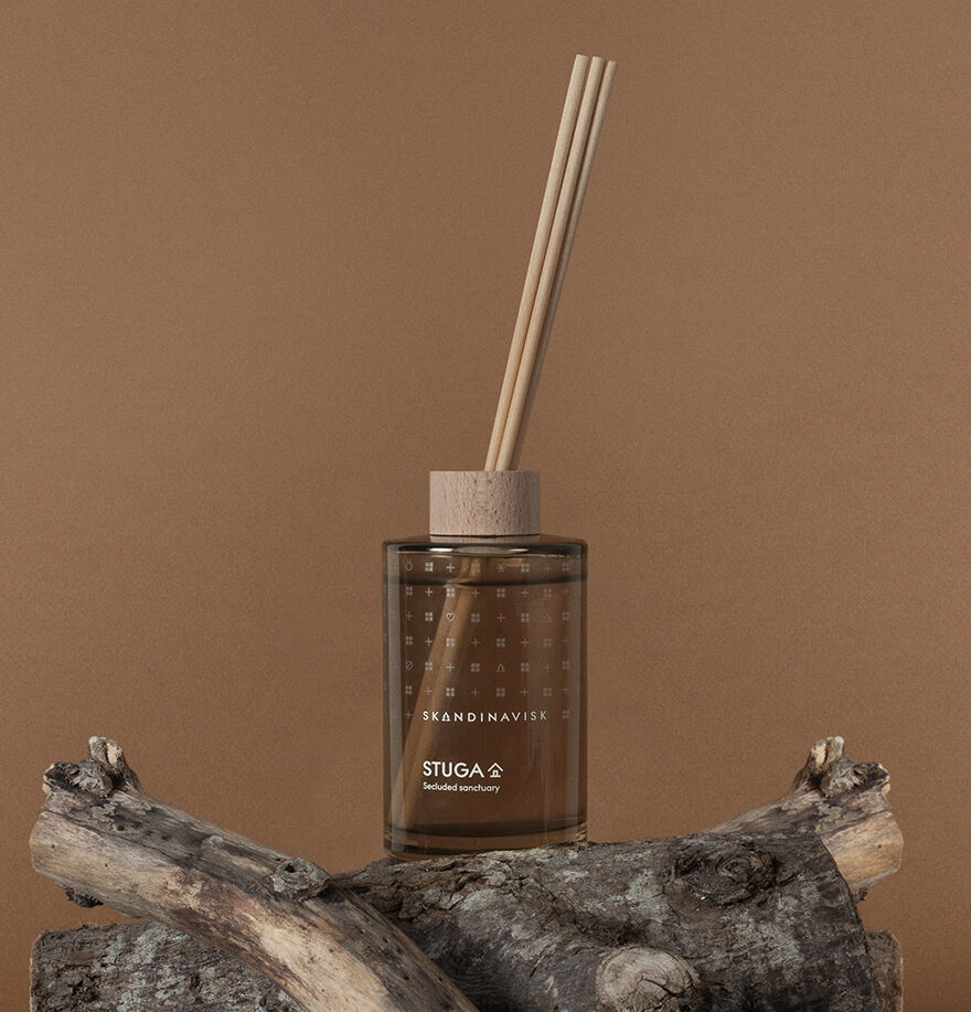 The Skandinavisk Scent Diffuser Seasonal Stuga, 200ml, with wooden sticks, sits on driftwood pieces and brings a refined room fragrance, set against a brown background.