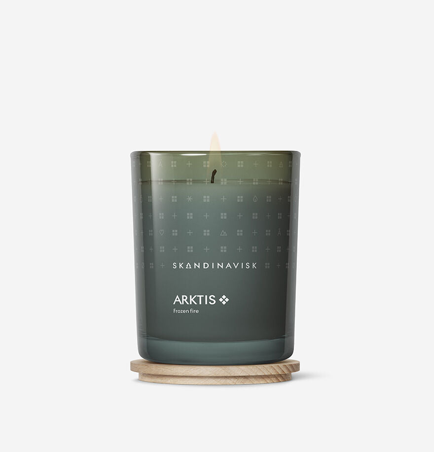 The Skandinavisk Scented Candle Arktis 50h, 200g, sits on a round wooden coaster against a white background, radiating fresh arctic sage fragrance in a sustainable green glass design.