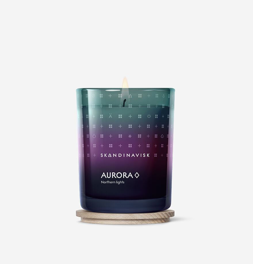 The lit Skandinavisk Seasonal Aurora Scented Candle 50h, 200g, made with Swedish rapeseed wax and set in a blue-purple gradient glass jar on a wooden coaster, is shown against a plain white background.