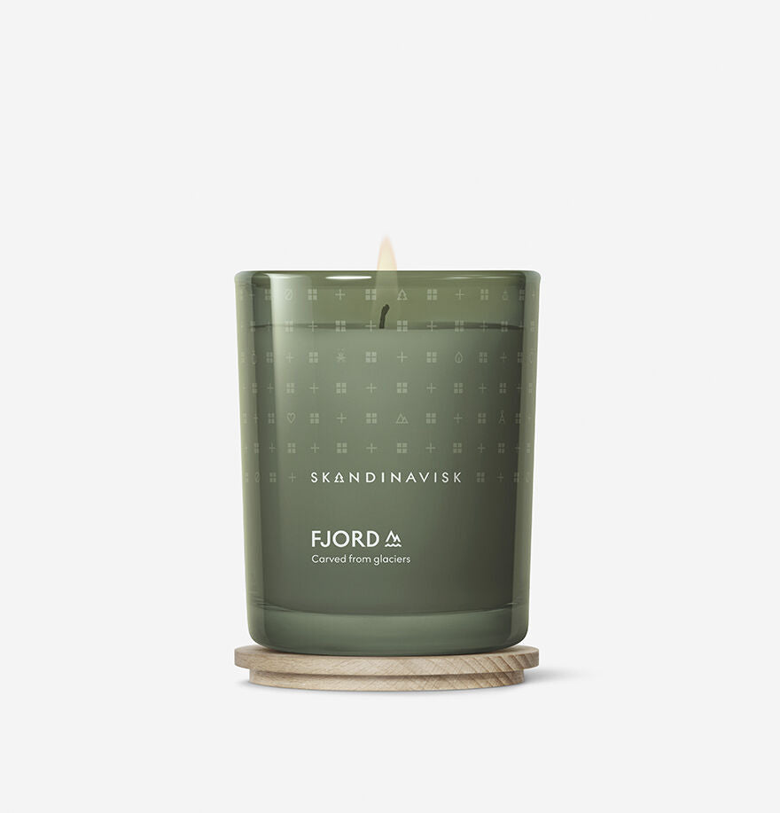 The Skandinavisk Scented Candle Fjord 50h, 200g, filled with Swedish rapeseed wax in green painted glass, sits on a round wooden coaster as its gentle flame glows warmly.