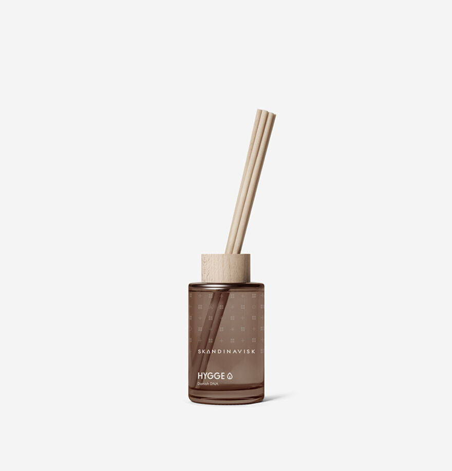 A 100 ml Skandinavisk Scent Diffuser Hygge (Small), alcohol-free, in a brown glass bottle with wooden cap and three reed sticks, evokes Danish coziness (hygge) and is set against a plain white background.