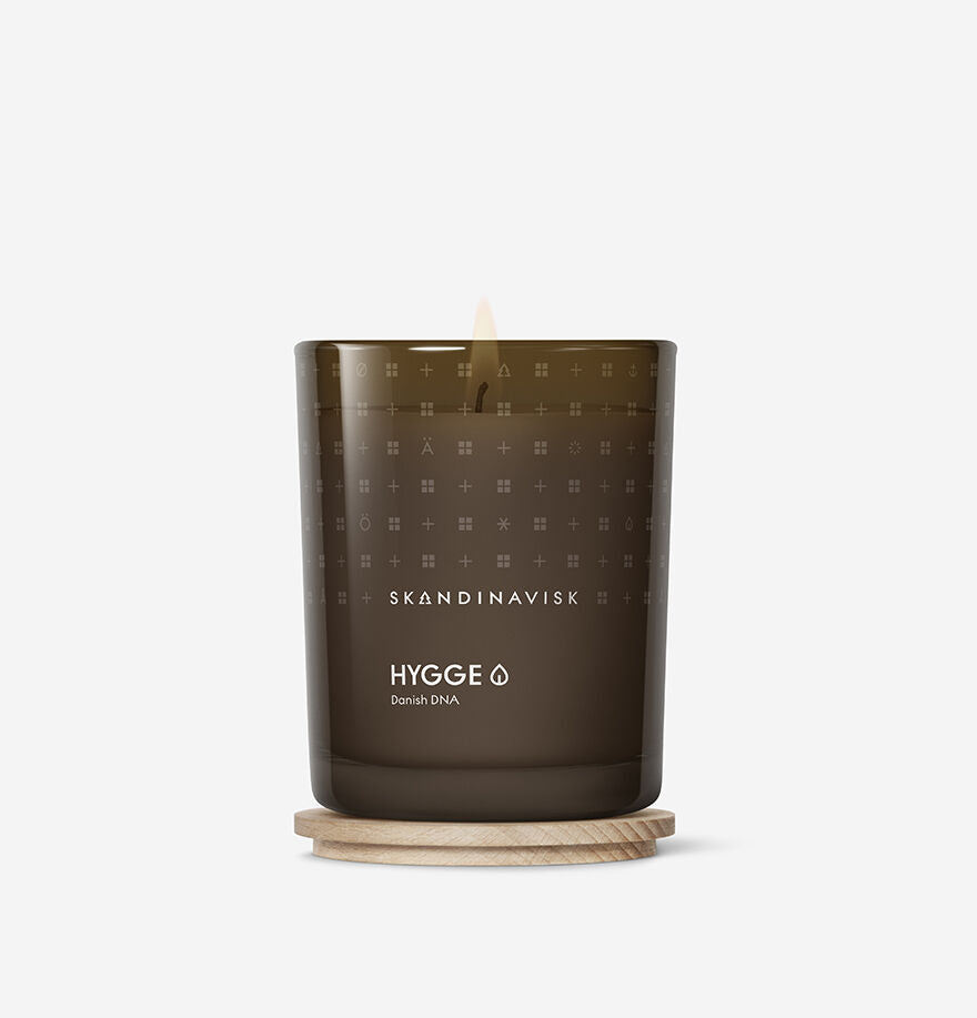 The Skandinavisk Scented Candle Hygge 50h, 200g, made from Swedish rapeseed wax, glows in a dark glass container placed on a round wooden coaster against a plain white background.
