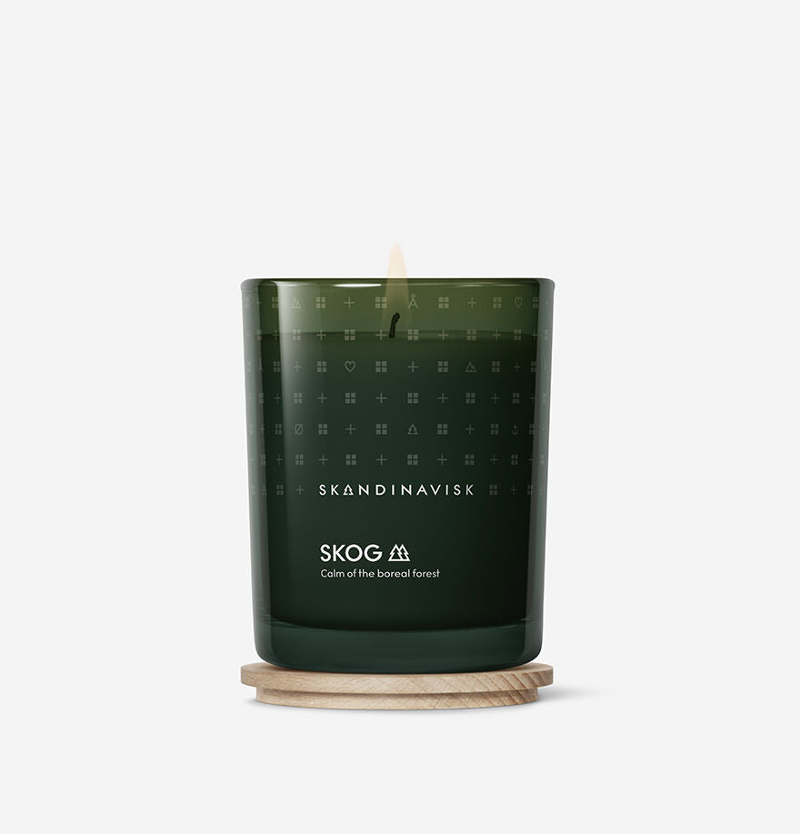 A green glass Scented Candle Skog 50h, 200g by Skandinavisk, made with Swedish rapeseed wax, burns on a round wooden coaster against a white background.