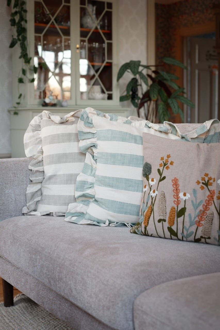 A light gray sofa features two striped ruffle-edged pillows and a Strömshaga Cushion Cover Elodie Embroidered Beige with Flowers (40 x 60 cm), set in a cozy living room with plants and a glass-front cabinet in the background.