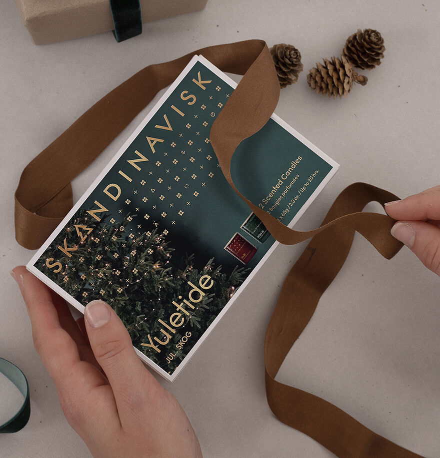 A person holds the Skandinavisk Giftset Yuletide—2 Scented Candles, Jul & Skog—partly wrapped with a brown ribbon, with pinecones and a gift nearby on a beige surface for a festive seasonal candle display.