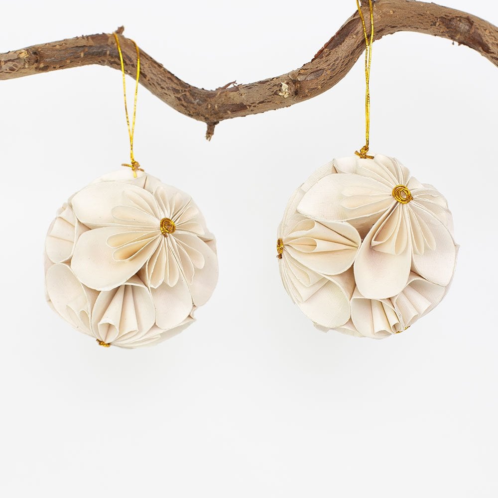 Two handmade Flower Ball Christmas Ornament 2-pack in White spherical ornaments made of folded white paper flowers hanging from a branch by gold strings, representing an ethical choice.