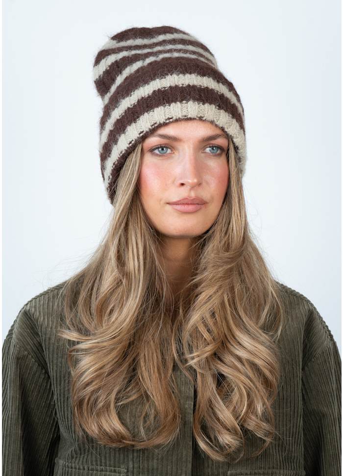 A woman with long, wavy blonde hair wears the Sui Ava Clara Beanie in Brown & Beige and an olive green corduroy jacket, looking straight ahead against a plain light background.