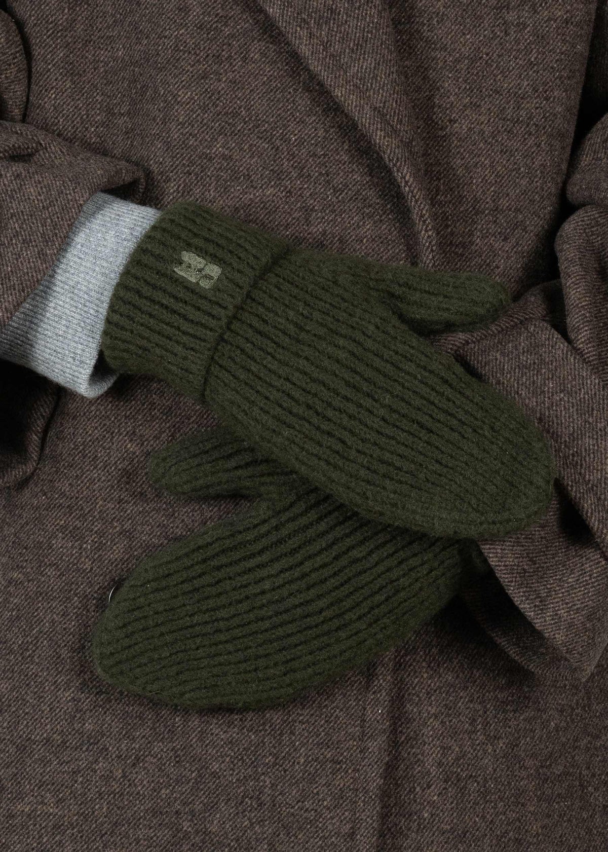 A person wearing Sui Ava Brenda Mittens in Dark Green, featuring a chunky rib-knit style and a small embroidered logo on the cuff, pairs them with a brown coat over a light gray sleeve.