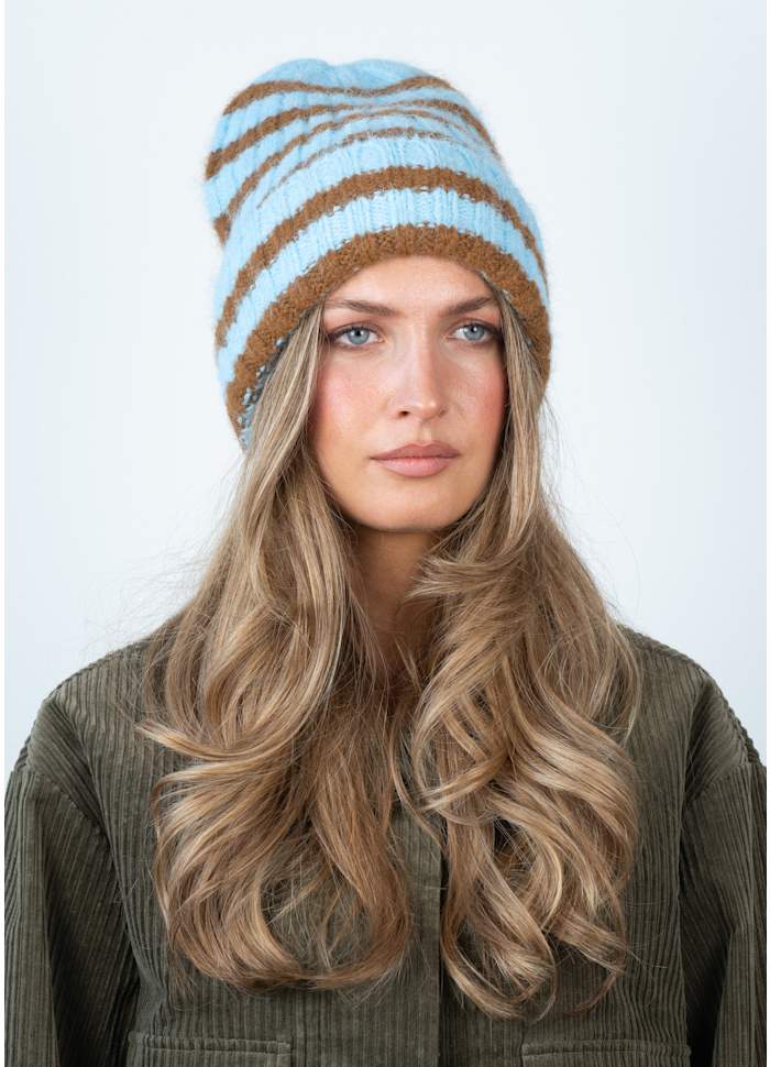A woman with long wavy blonde hair wears the Sui Ava Clara Beanie in Blue and Khaki with a green corduroy jacket, facing forward against a plain light background.