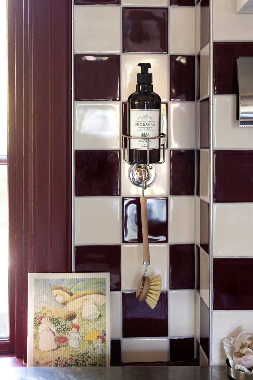 A soap dispenser and brush hang on a square-tiled wall with maroon and cream tiles; on the counter below sits a painting of fairies and flowers inspired by Elsa Beskow, accompanied by neatly folded Strömshaga Dishcloths featuring the “Elsa Beskow Children of the Forest” design.