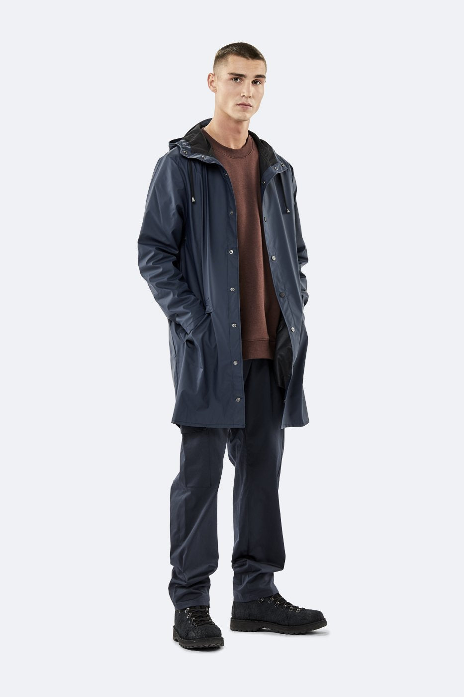 A man faces forward wearing the Rains Unisex Long Jacket in Navy Blue by Rains, paired with a brown shirt, dark pants, and black shoes against a plain light background.