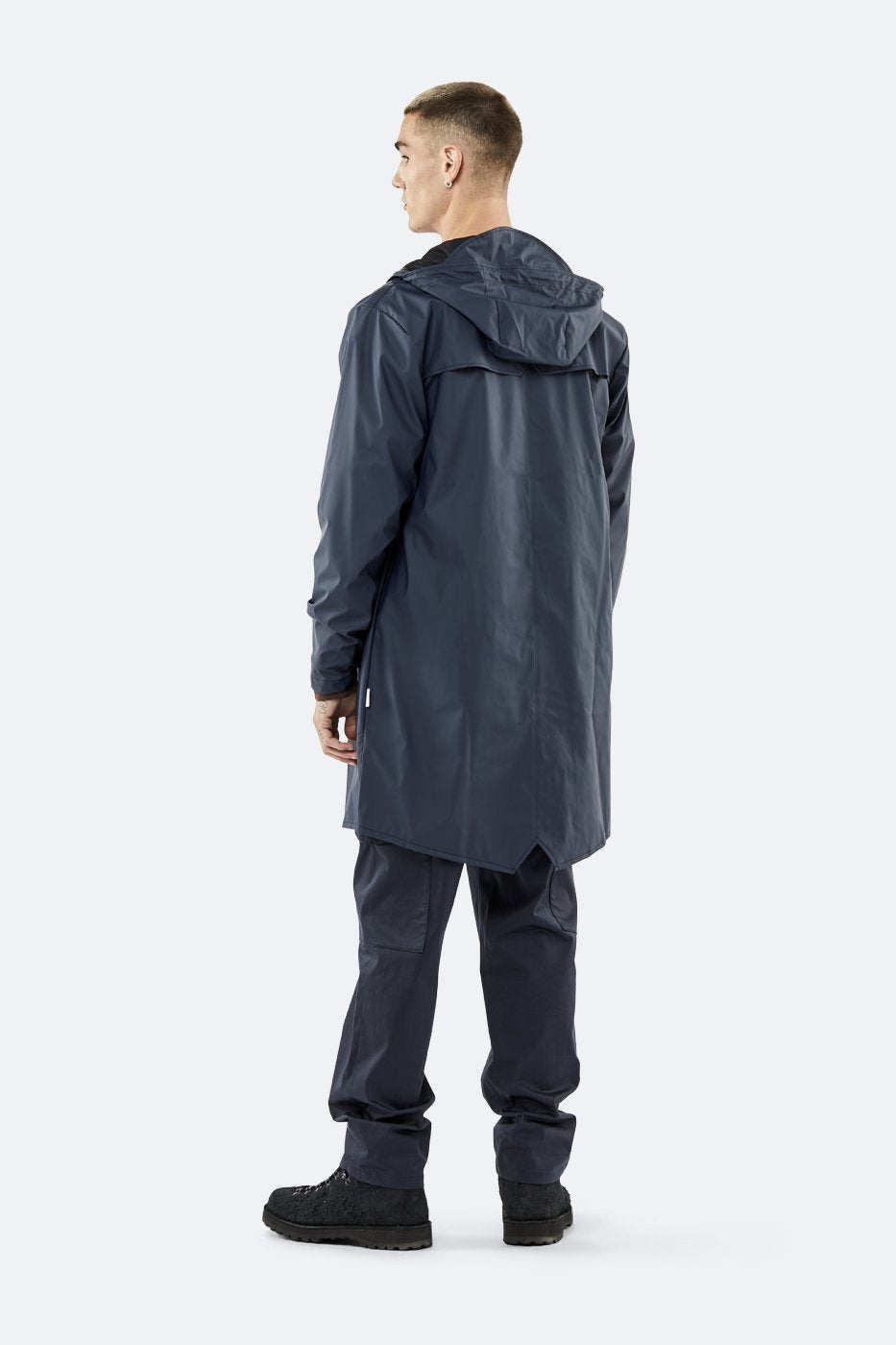 A person faces away, wearing the Rains Unisex Long Jacket in Navy Blue by Rains, paired with matching dark blue pants and black shoes, set against a plain light background.