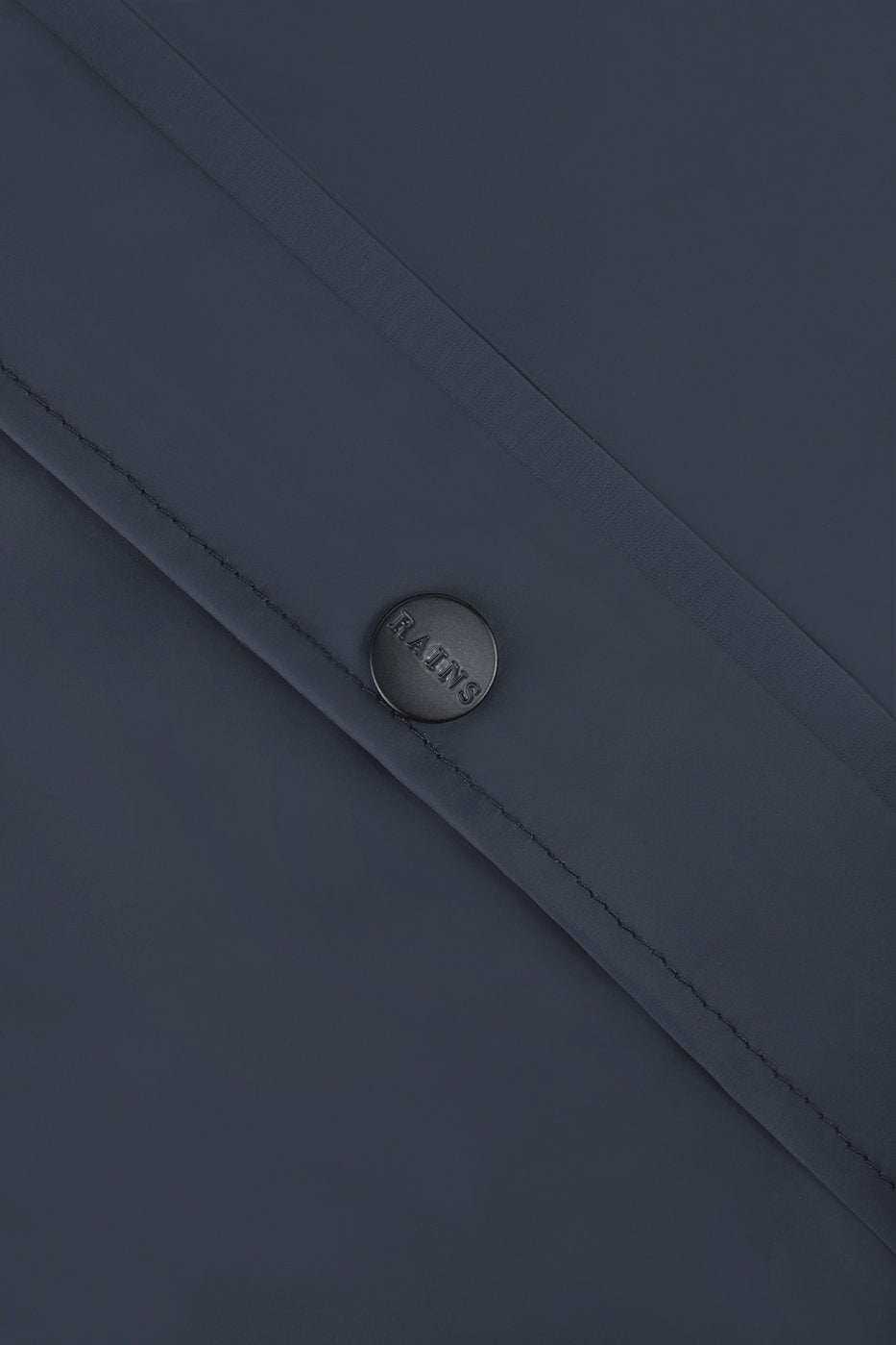 Close-up of the Rains Unisex Long Jacket in Navy Blue, showing a metallic Rains-embossed button secured to a flap with visible stitching.