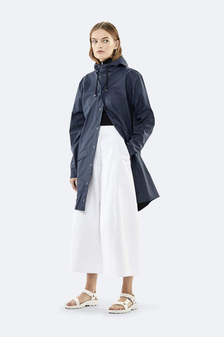 A woman wears the Rains Unisex Long Jacket in Navy Blue by Rains, styled with a black top, white wide-leg pants, and white sandals against a plain light background.