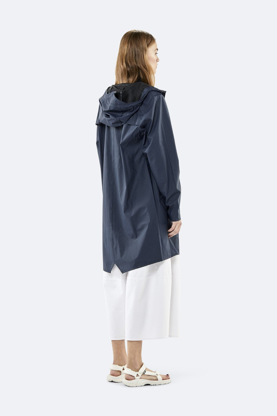 A person stands sideways against a plain light background, wearing the Rains Unisex Long Jacket in Navy Blue by Rains, paired with white wide-leg pants and white sandals.