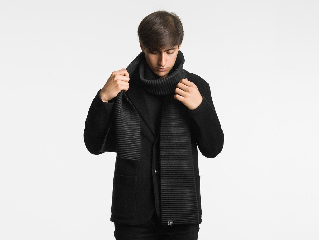 A person in a black coat and pants adjusts the Design House Stockholm Pleece Scarf Long in Black around their neck against a plain white background.