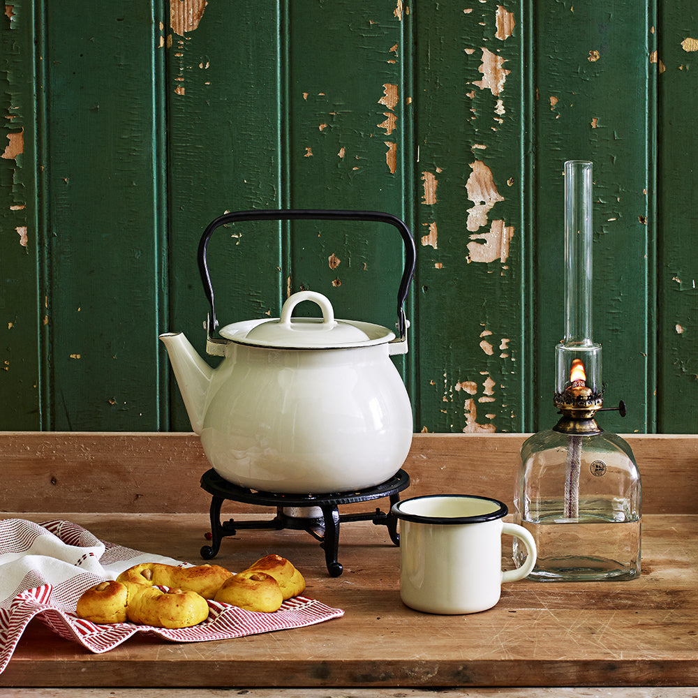A white teapot on a stand, a matching mug, and pastries sit beside the Strömshaga Kerosene Lamp Elisabeth in Glass, Square with Antique Brass 33cm on a wooden table, set against a green, weathered wooden wall.