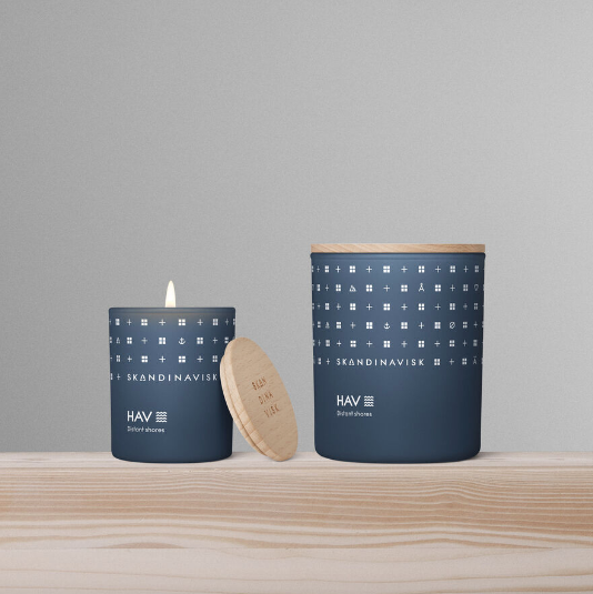 Two blue Skandinavisk Mini Scented Candle Hav 20h, 65g candles made with Swedish rapeseed wax rest on a light wood surface before a gray backdrop. One is lit, the other lidded; both capture the fresh scent of Scandinavian sea air.