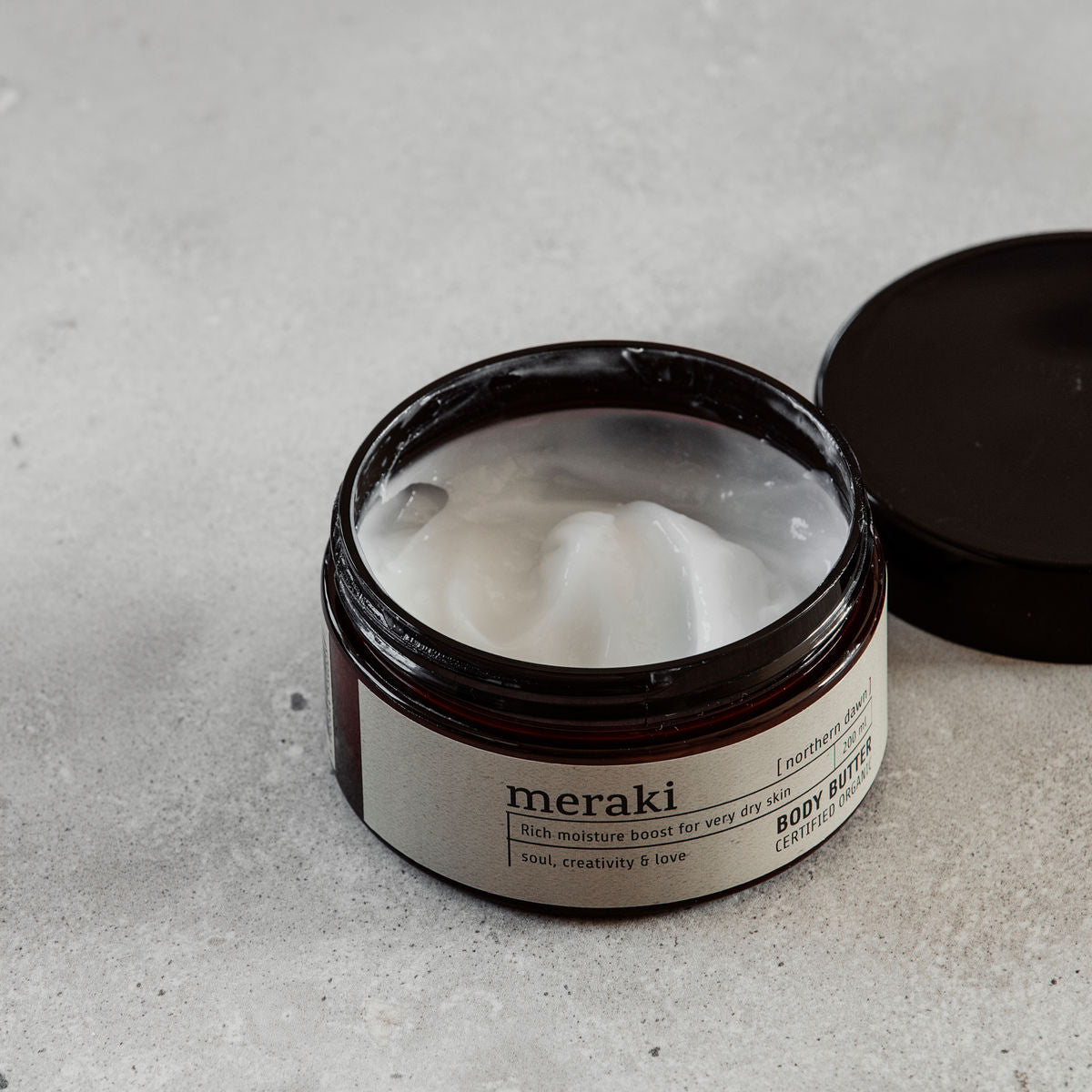 An open jar of Meraki Body Butter Organic, Northern Dawn, with its black lid beside it, reveals creamy white moisturizer inside on a gray surface.