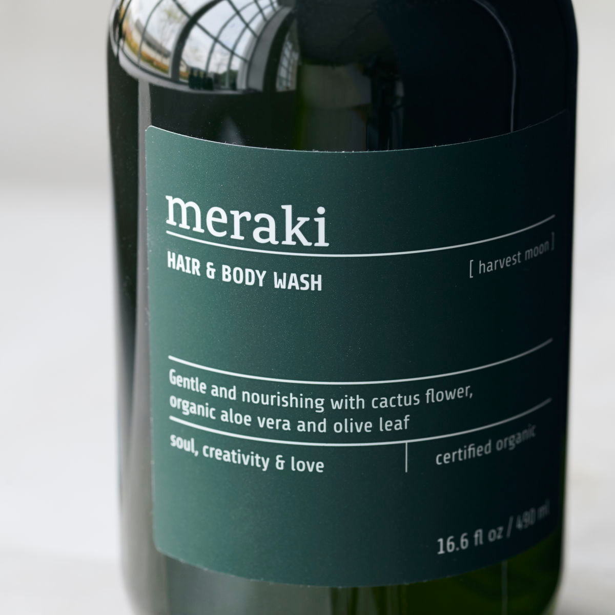 A dark green bottle labeled Meraki Hair & Body Wash, Harvest Moon, features hydrating ingredients like cactus flower, aloe vera, and olive leaf for nourishing hair and skin.