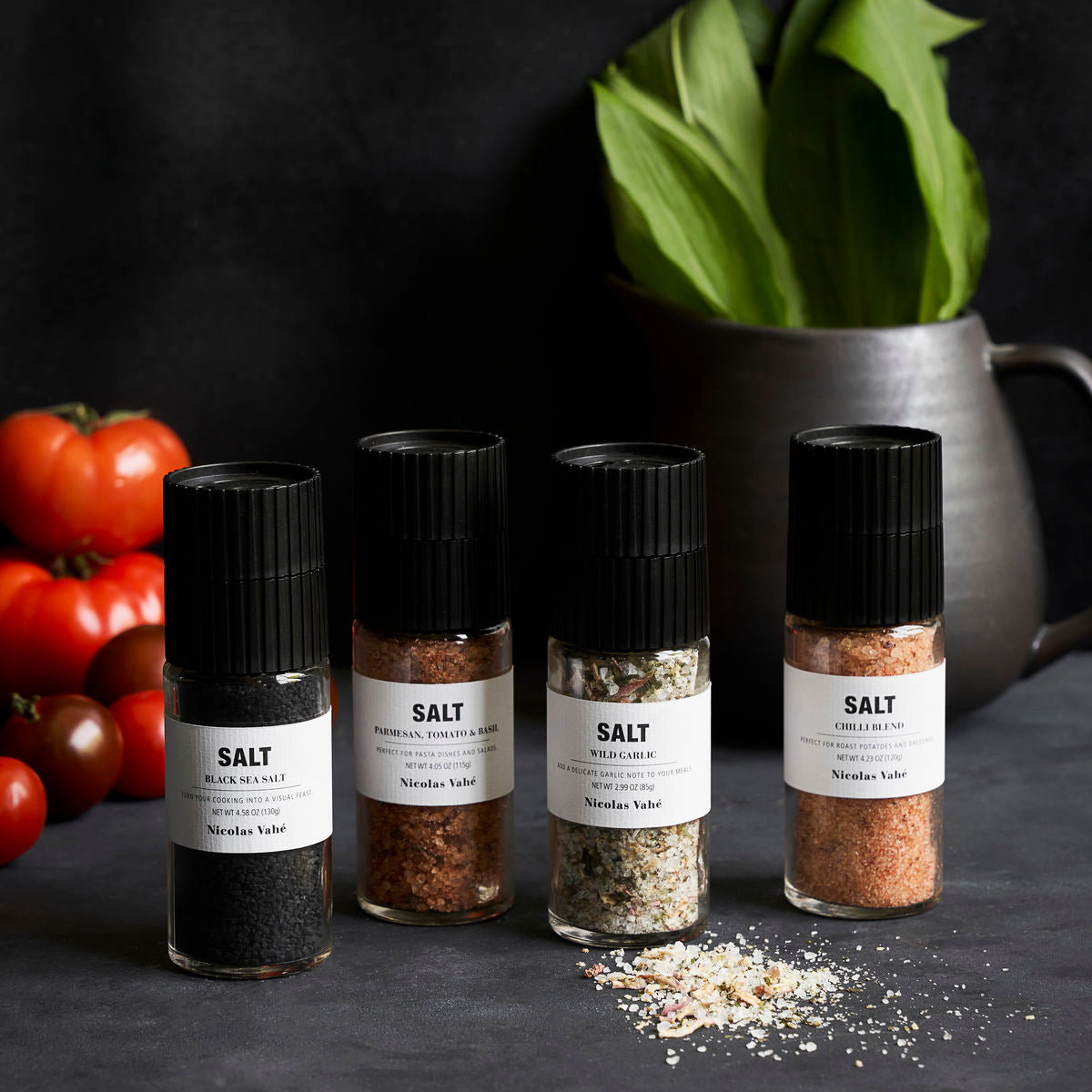 Four ceramic grinders labeled Salt from the Nicolas Vahé Gift Box Savoury Salts—featuring Parmesan, Tomato & Basil, Black Salt, Wild Garlic, and Chili—are displayed on a dark surface with tomatoes and a vase of green leaves behind them. Salt is scattered in front.