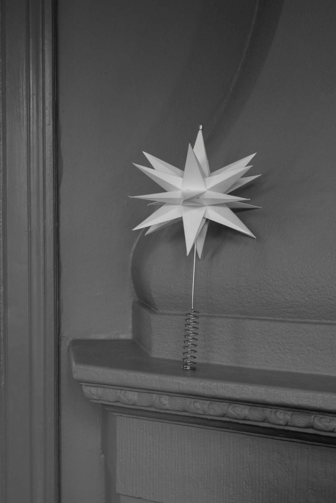 The Watt & Veke Topstar Sputnik, handmade in paper (25cm), sits on a mantel as a modern accent or Christmas tree topper, its white star and spring base contrasting stylishly against a dark, smooth wall.
