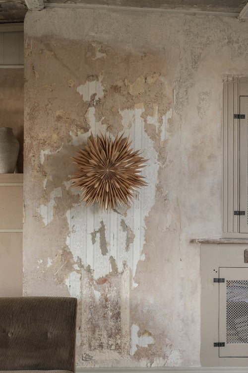 A textured beige wall with peeling paint displays the Star Maja in Paper, Gold 60cm by Watt & Veke. A brown chair and part of a beige cabinet are also visible.