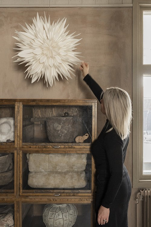 A woman in a black dress touches the Watt & Veke Star Maja in Paper, White 60cm, displayed above a wooden cabinet filled with folded textiles and baskets.