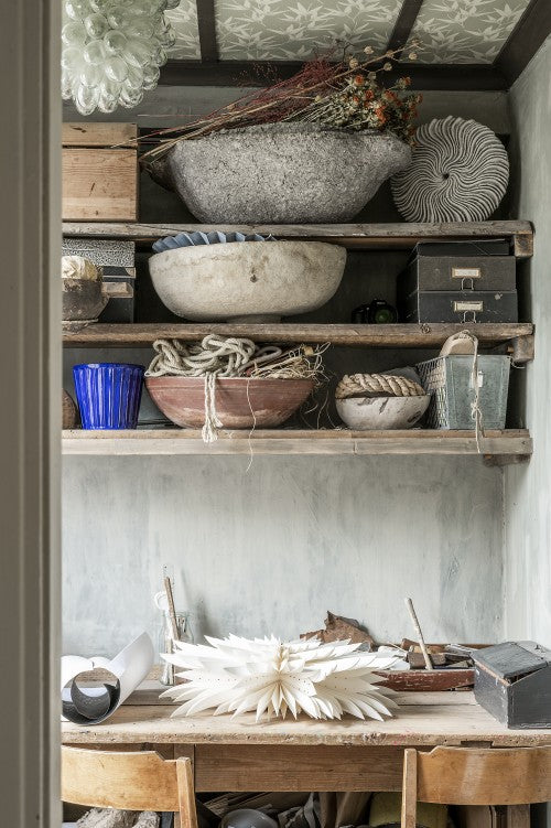 A small workspace with wooden shelves holds bowls, rope, boxes, and dried flowers; on the table sits Watt & Veke’s Star Maja in Paper, White 60cm in Box. Two wooden chairs are pushed under the table.