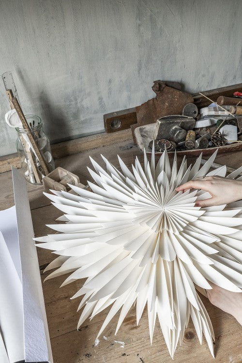 A person assembles a Watt & Veke Star Maja in Paper, White 60cm in Box on a wooden table, with tools and supplies visible in the background.