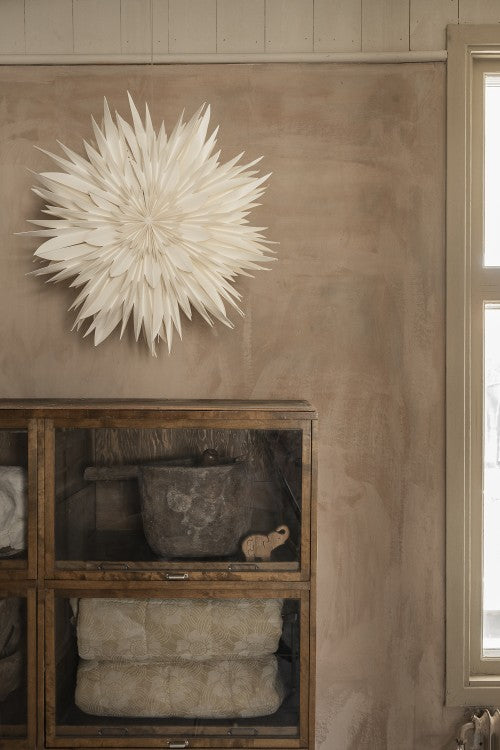 A Watt & Veke Star Maja in Paper, White 60cm, hangs elegantly on a beige wall above a glass-front cabinet holding folded blankets, a stone pot, and a small carved animal.
