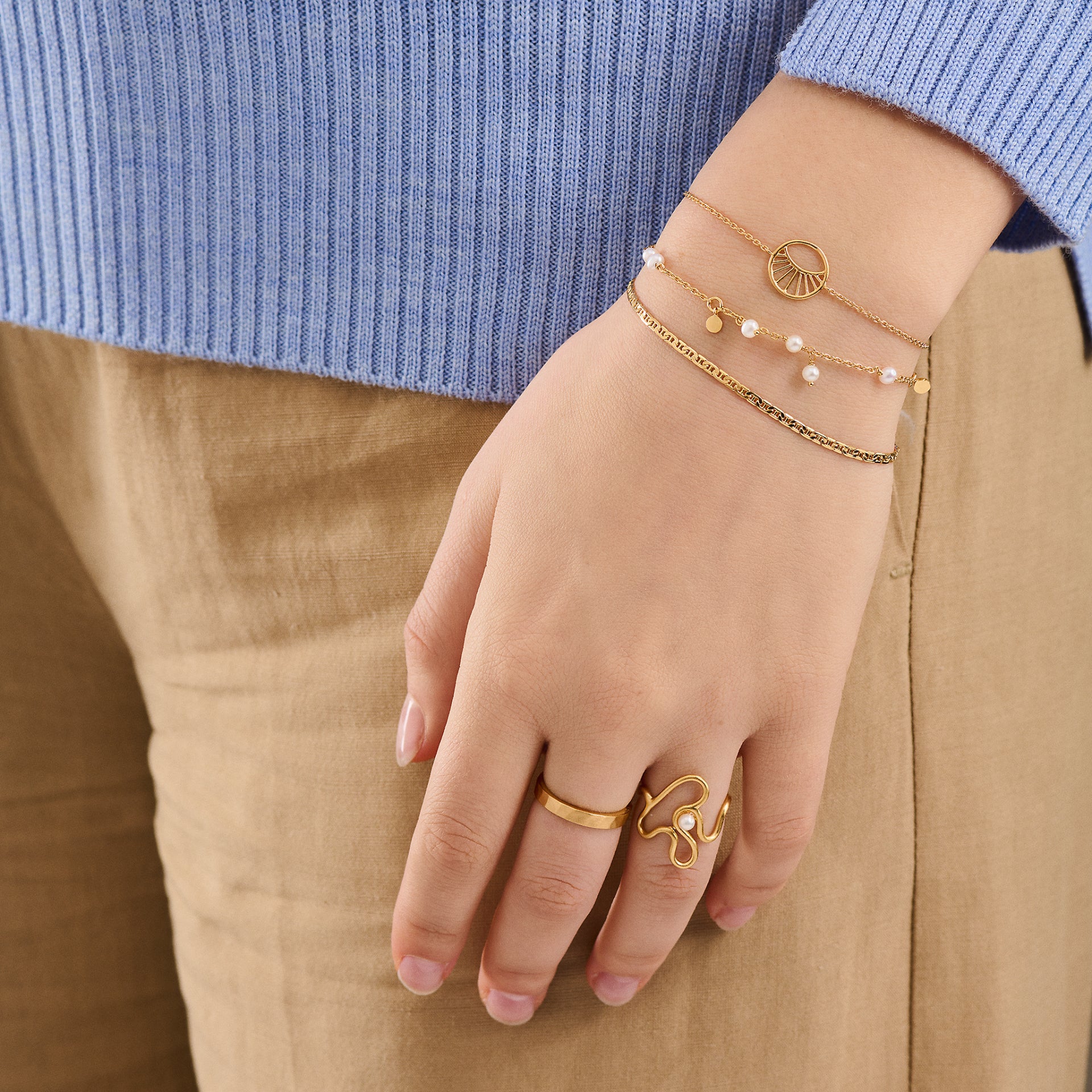 A hand adorned with Pernille Corydon’s Ocean Pearl Bracelet in Gold, plus gold rings and two delicate bracelets, is shown against a blue sweater and beige pants.