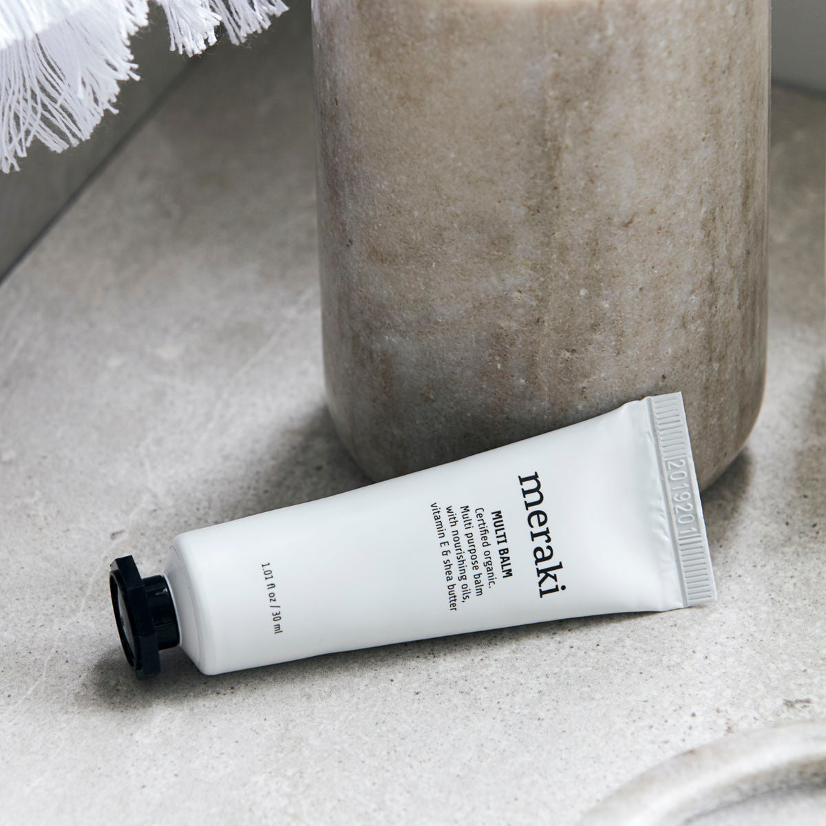 A 30 ml tube of Meraki Organic Multi Balm for Moisturising Dry & Damaged Skin sits on a gray stone beside a beige cylindrical container—ideal for soothing dryness.