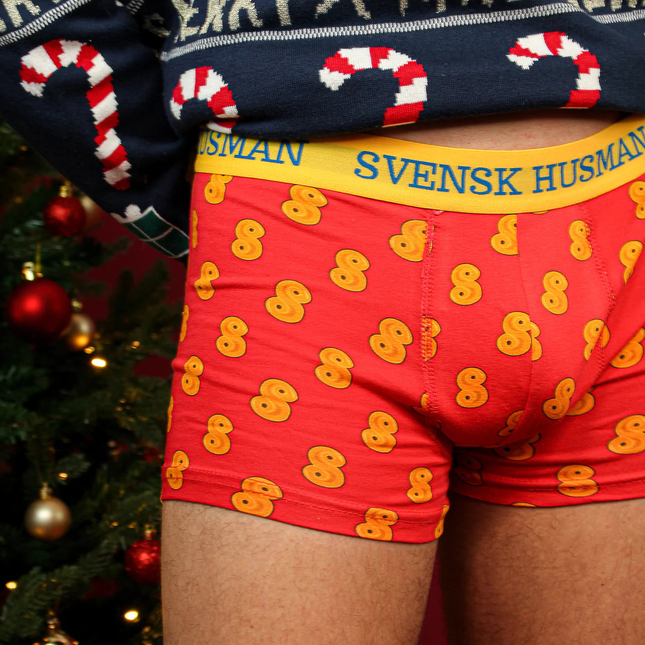 A person in red Lussebulle (Saffron Bun) Boxershorts by Svensk Husman Socks, featuring yellow patterns and SVENSK HUSMAN on the waistband, stands next to a decorated Christmas tree.