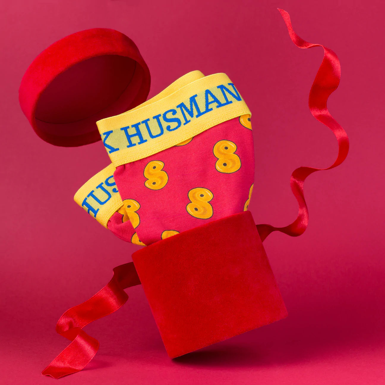 A pair of Lussebulle (Saffron Bun) Boxershorts by Svensk Husman Socks, in red and yellow cotton with HUSBAND on the waistband, is presented in an open red velvet box with a matching ribbon against a red background.