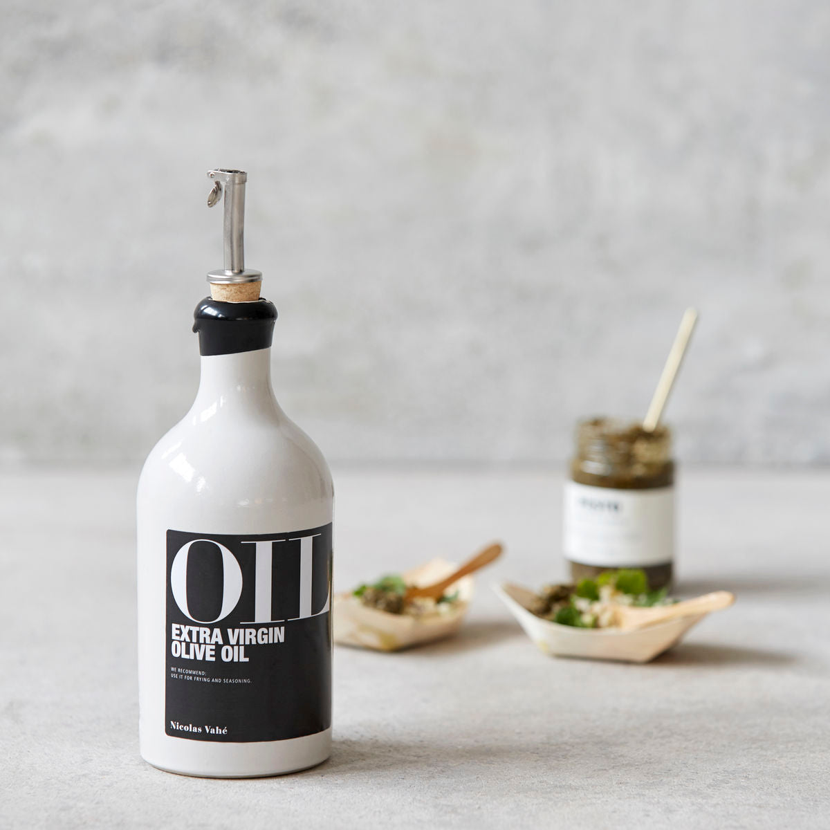 A House Doctor Pourer for Oil & Bottles, small, stands in the foreground, while a jar and two small dishes of food are blurred in the background.