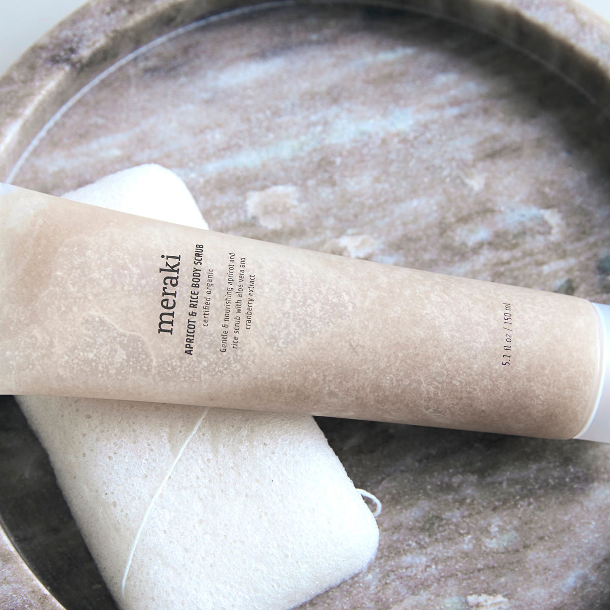 A beige tube of Meraki Apricot & Rice Body Scrub with organic aloe vera rests on a white sponge atop a round stone dish.