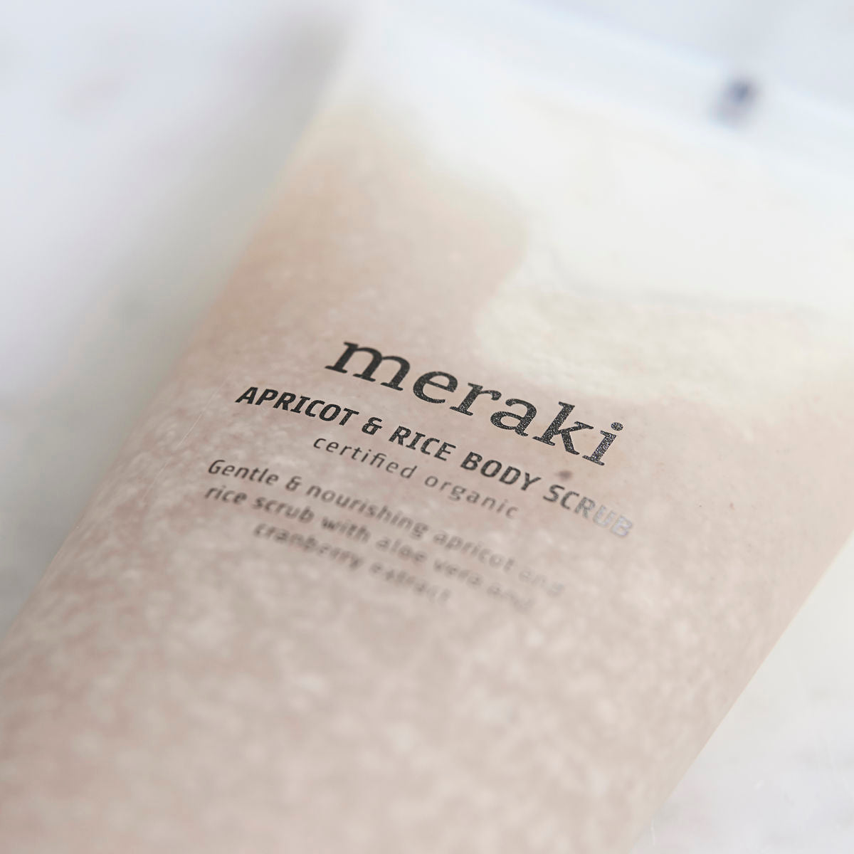 A close-up of a Meraki Apricot & Rice Body Scrub w. Aloe Vera, Organic highlights its label and showcases the creamy, grainy texture with exfoliating granules inside the transparent packaging.