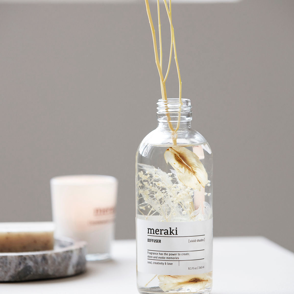 The Meraki Diffuser, Vivid Shades, features a clear glass bottle filled with dried flowers and reeds, placed on a white surface to create a calming vibe, with a blurred candle and soap in the background.