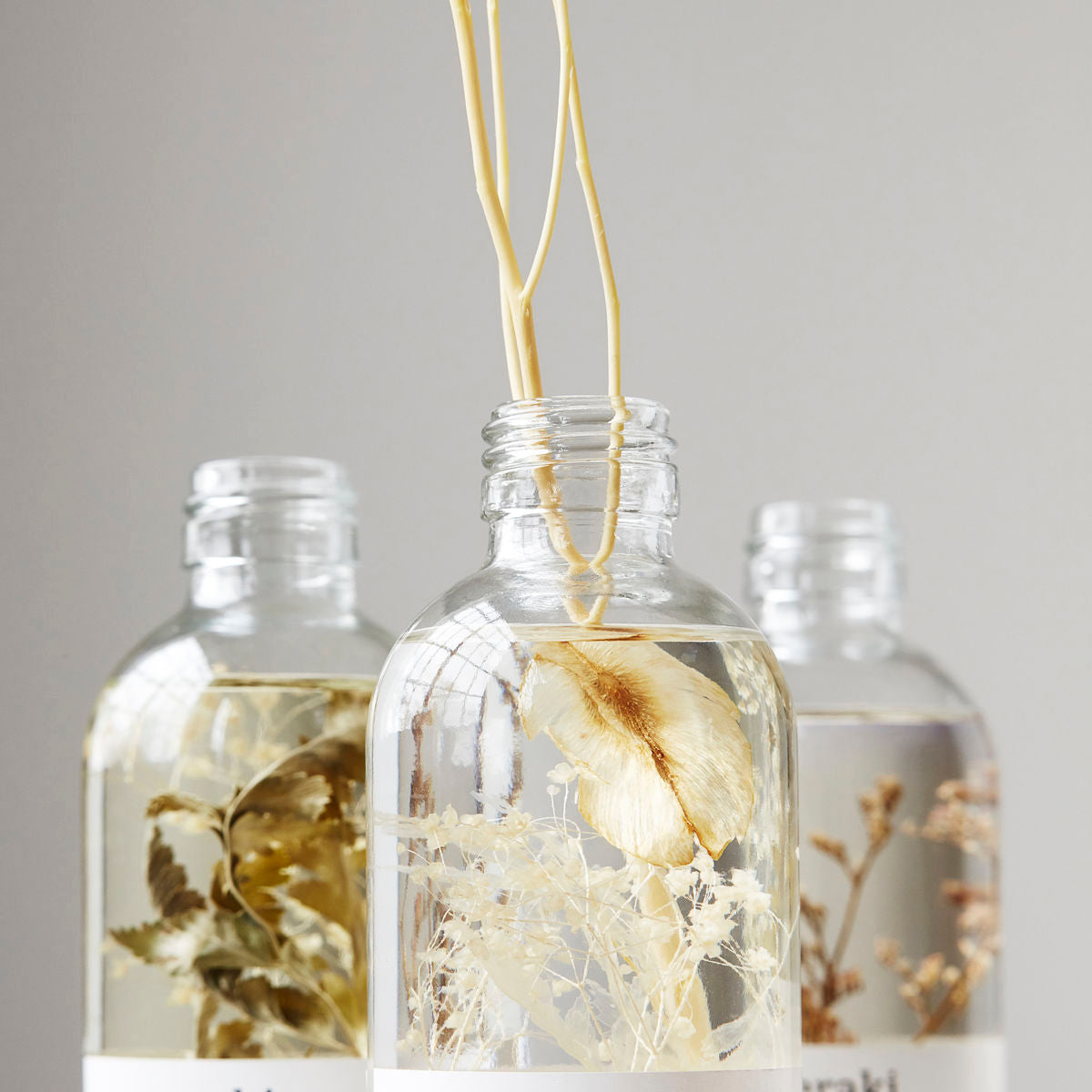 Three clear glass bottles filled with liquid and dried botanicals are arranged together against a plain background, evoking the soothing vibe of the Meraki Diffuser, Vivid Shades.