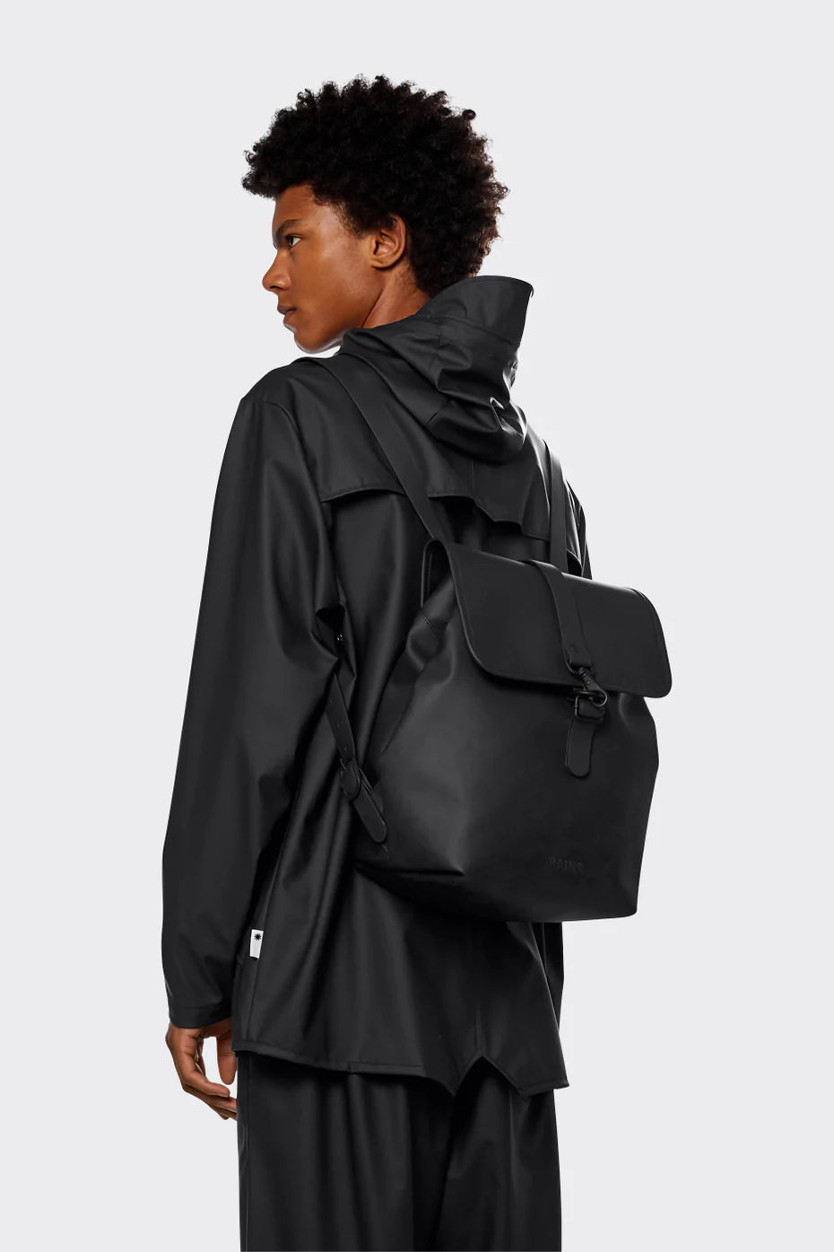 A person in black rain jacket and pants, seen from the side, carries the Rains Bucket Backpack in Black by Rains with a flap closure against a plain light background.