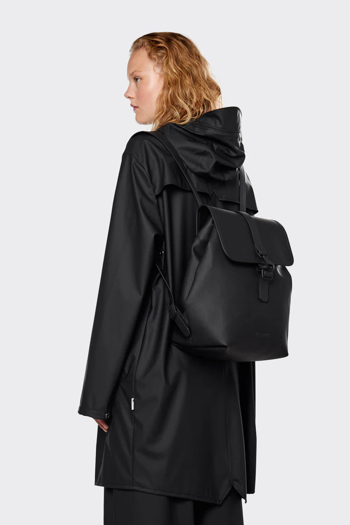 A person stands sideways against a plain light gray background, wearing a black raincoat and carrying the Rains Bucket Backpack in Black by Rains, made from weatherproof PU fabric.