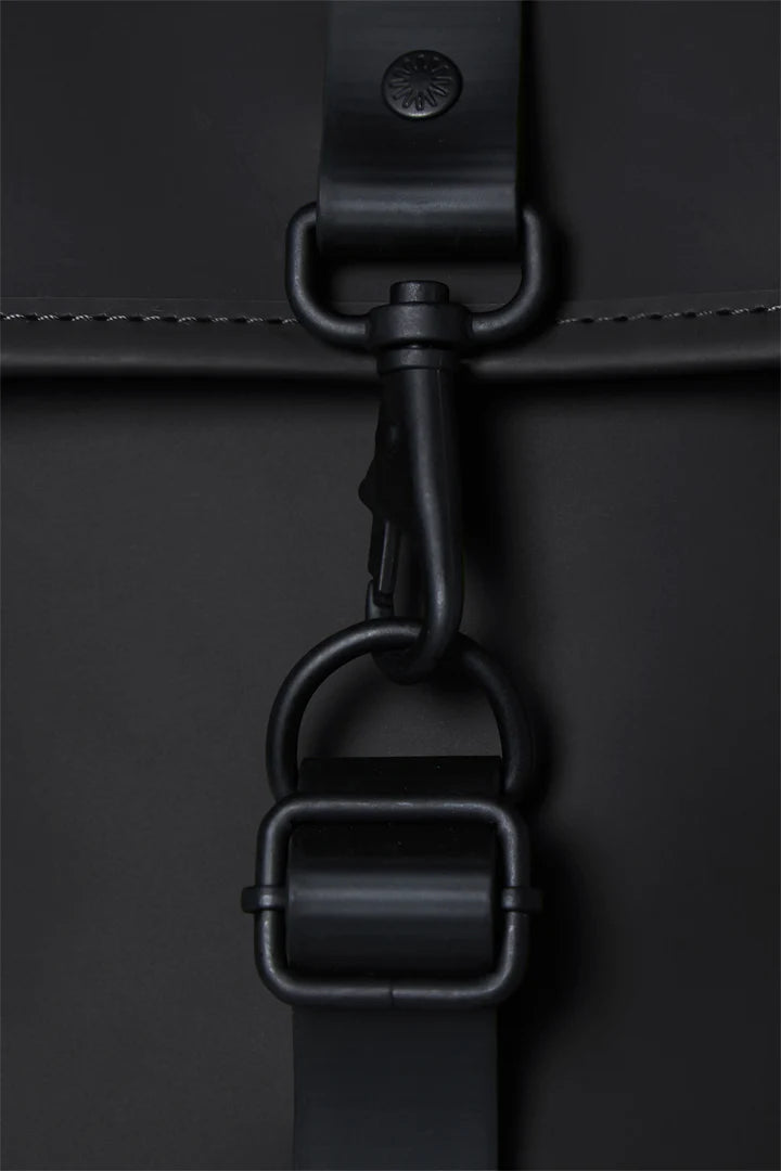 Close-up of the black metal clasp and buckle on the Rains Bucket Backpack in Black by Rains, showing detailed stitching, a leather strap, and durable PU fabric.