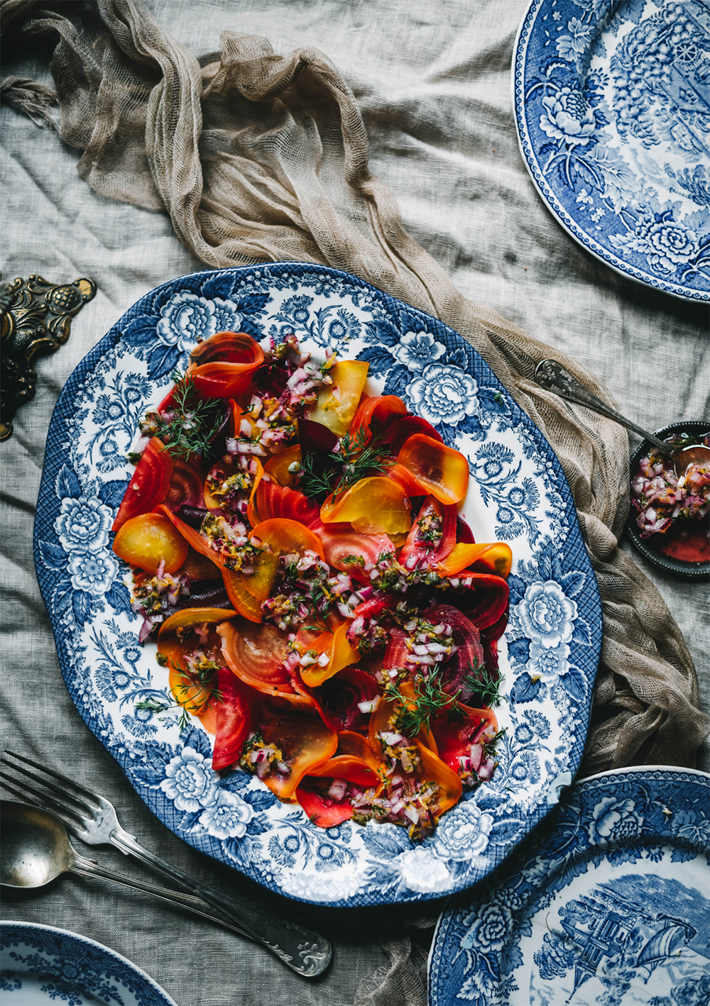 A vibrant beet and carrot salad, featured in Cozy Publishings Nordic Winter - Cook Book, is served with onion and herb garnish on a blue-and-white floral plate over linen, capturing the essence of classic Finnish Christmas cuisine.