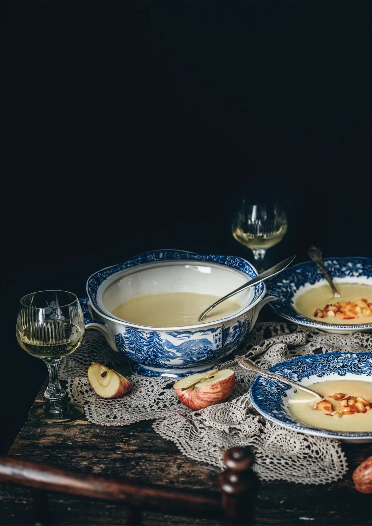 The Nordic Winter - Cook Book by Cozy Publishing rests on a wooden table with a lace cloth, surrounded by apple slices, soup bowls, and white wine—setting the scene for savoring Finnish Christmas cuisine.