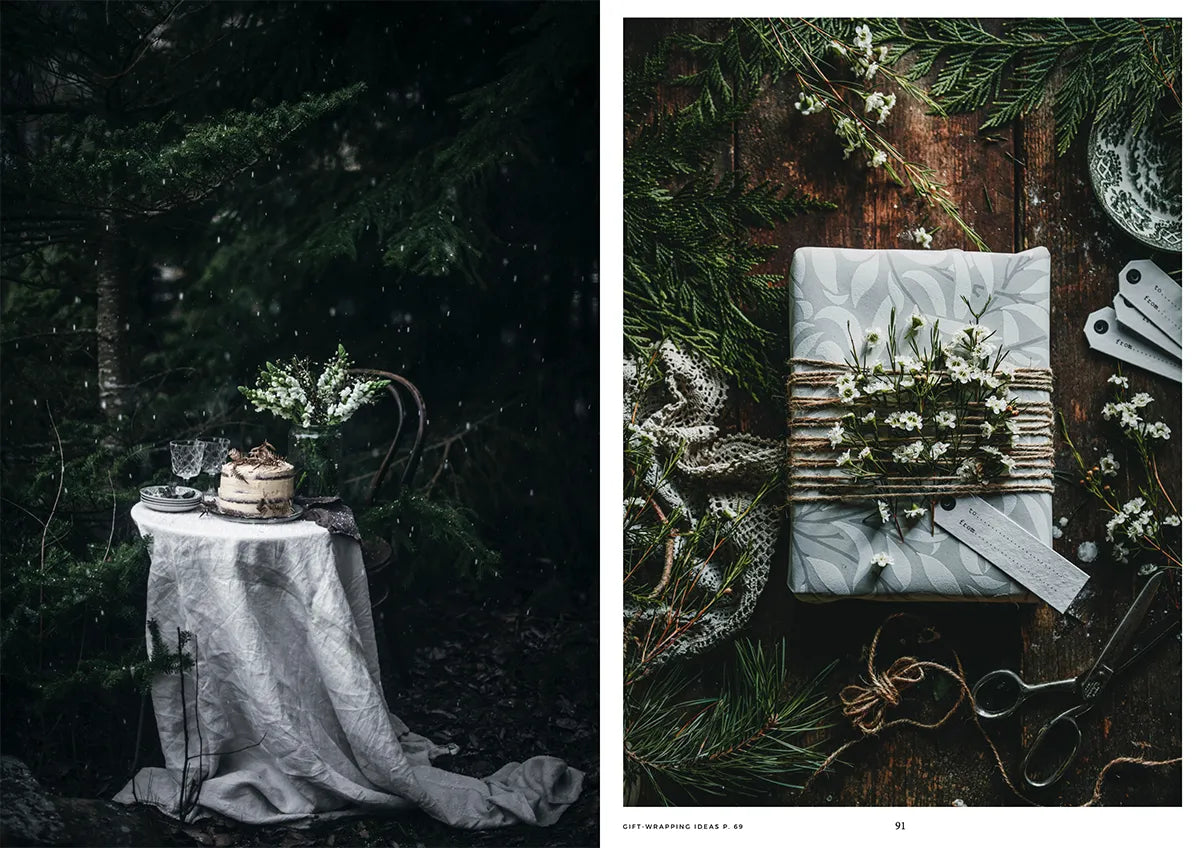 A small table with cake, glasses, and flowers stands in a dark forest; nearby, Cozy Publishing’s “Nordic Winter – Cook Book” evokes rustic Nordic desserts under the trees, wrapped with twine and greenery, scissors resting atop.