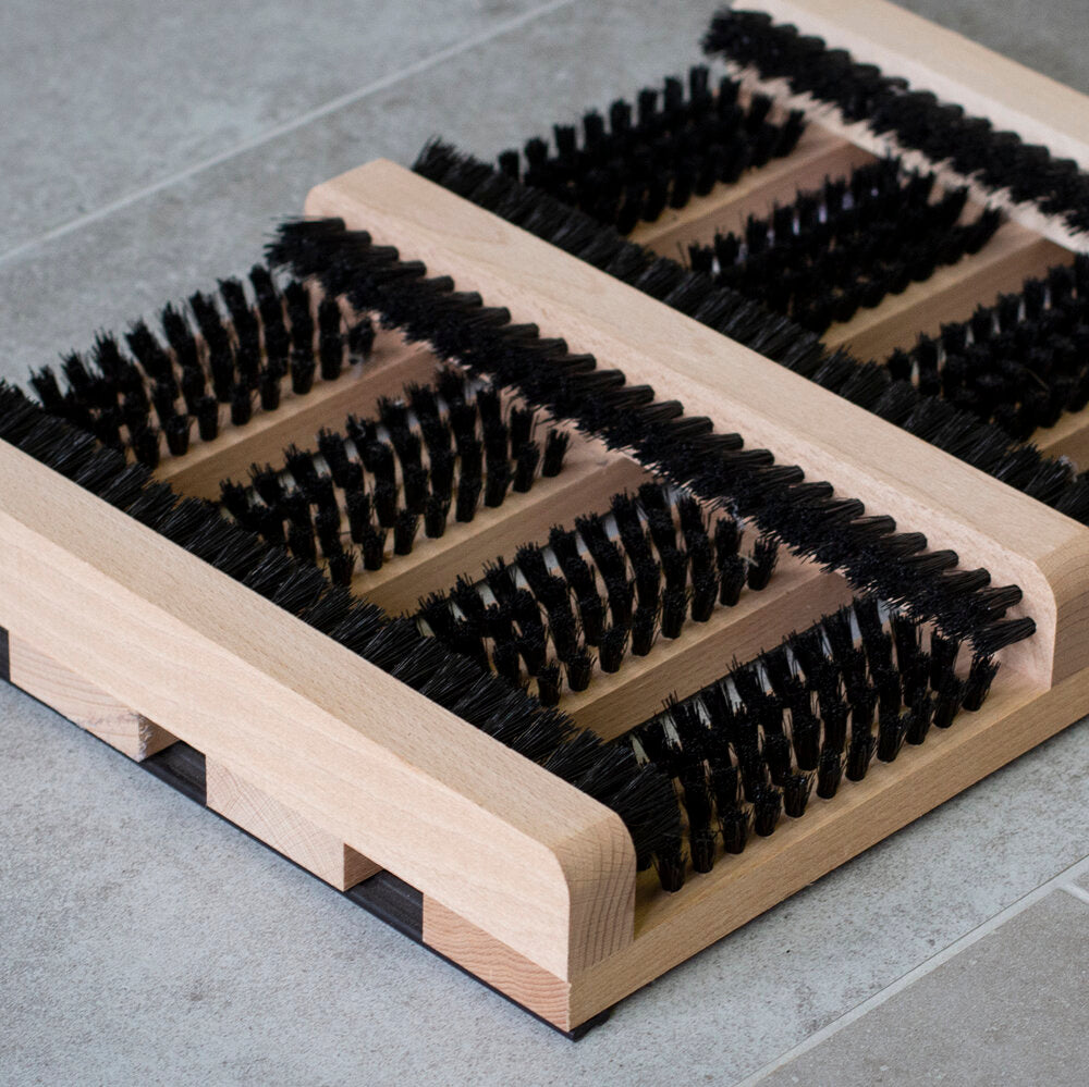 The Iris Hantverk Bootscraper in Beech Wood (36x27cm) features multiple rows of stiff black bristles and is ideal for outdoor use; shown here on a gray tiled floor.