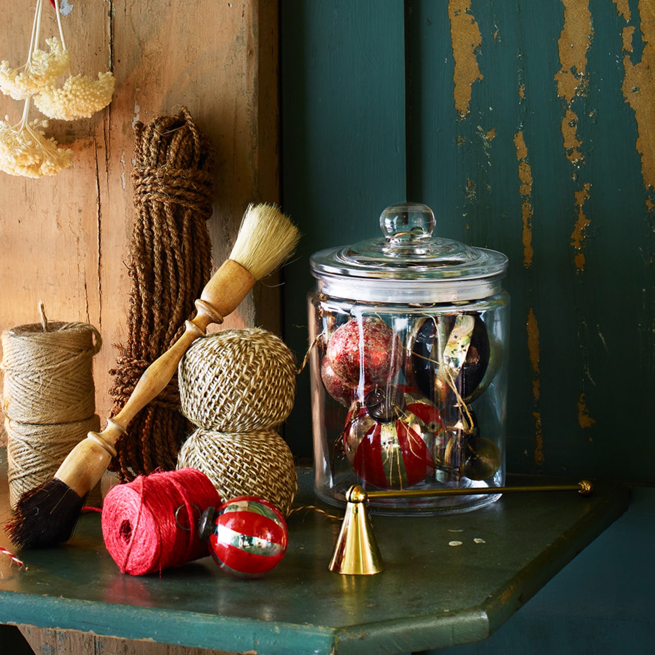 A glass jar of Christmas ornaments sits on a table beside twine spools, red string, brushes, a small gold bell, and the Strömshaga Candle Snuffer in Brass.