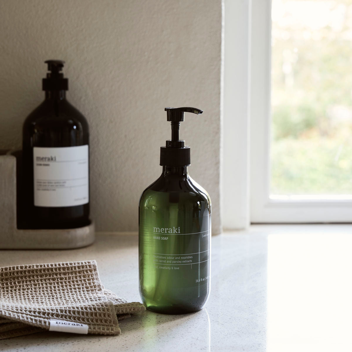 A green bottle of Meraki Hand Soap, Anti-Odour with a pump is on the countertop beside a folded beige towel, while a black bottle rests by the window. This soap neutralises food odours for fresh, clean hands.