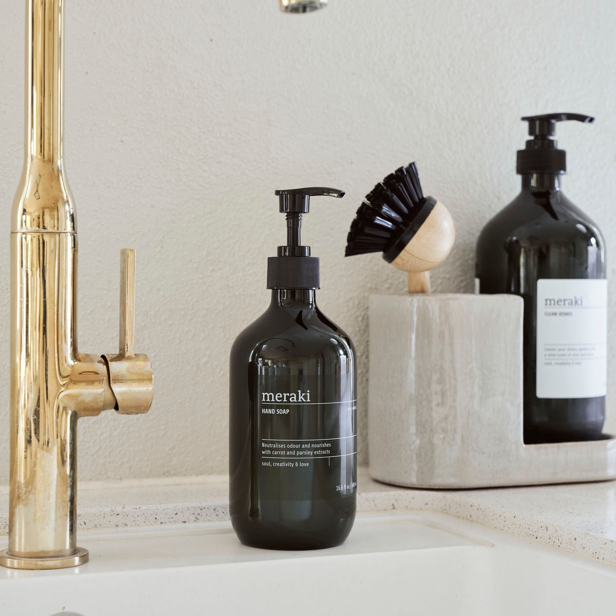 A stylish black glass Meraki Hand Soap, Anti-Odour bottle sits by a gold faucet on a white countertop, with a scrub brush and another bottle nearby. This soap effortlessly neutralises food odours from your hands.